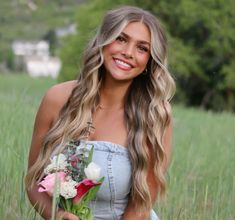 a beautiful young woman holding a bouquet of flowers in a grassy field with trees behind her