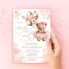 a hand holding up a pink and gold teddy bear baby shower card on a pink background