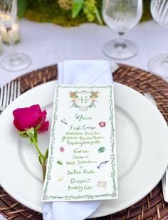 a white plate topped with a pink rose next to a wine glass and menu card