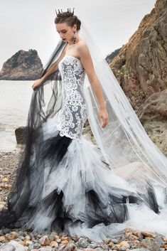 a woman in a wedding dress standing on rocks near the ocean with her veil flowing