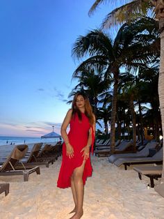 a woman in a red dress is standing on the beach near some lounge chairs and palm trees