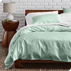 a bed with green sheets and pillows in front of a white brick wall next to a night stand