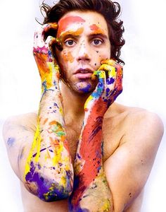 a man with his hands to his face covered in colorful paint and holding two fingers up to his face