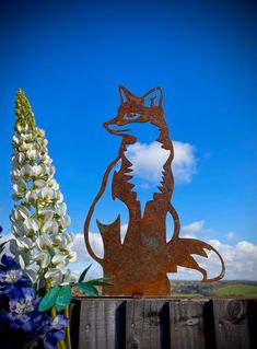 a metal fox sitting on top of a wooden fence next to flowers and a blue sky