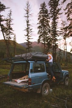 a man standing on the back of a blue jeep with its roof open and luggage strapped to it