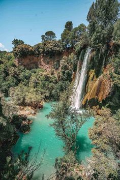 a waterfall in the middle of a forest with blue water and trees on either side