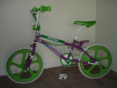 a purple and green bike sitting in front of a white wall next to a brown carpeted floor