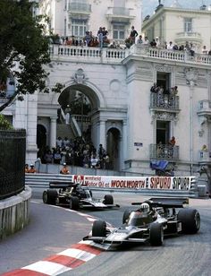 two racing cars driving down a race track in front of a white building with people watching