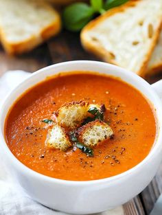 a bowl of tomato soup with croutons on the side and bread in the background