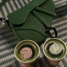 two cups of coffee sit on a table next to a green handbag and purse