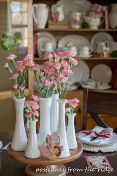 four white vases with pink flowers in them on a wooden table next to plates and dishes