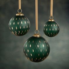 three green ornaments hanging from strings against a dark background with white dots on them and gold trimmings