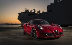 a red sports car parked in front of a large ship at sunset with the sun setting