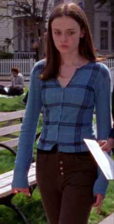 a woman walking down a sidewalk next to a park bench with a book in her hand