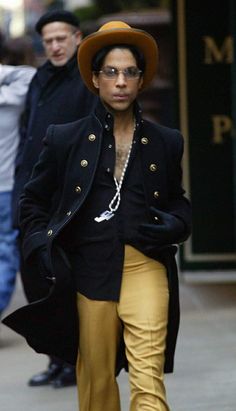 a man in yellow pants and a black jacket is walking down the street with a hat on