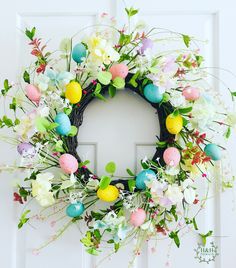 an easter wreath with eggs and flowers hanging on a white front door, decorated with greenery