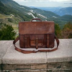 a brown leather briefcase sitting on top of a stone wall with mountains in the background