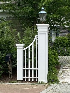 a white gate with a light on top in front of some bushes and trees near a house