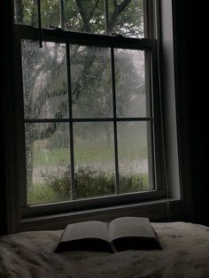 an open book sitting on top of a bed in front of a window covered in rain