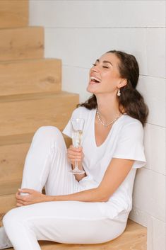 a woman sitting on the steps holding a glass of wine and laughing with her eyes closed