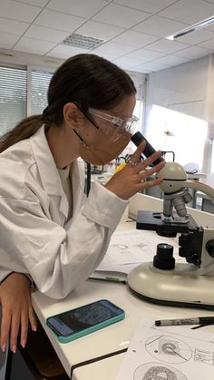 a woman in white lab coat looking through a microscope while holding a cell phone up to her ear