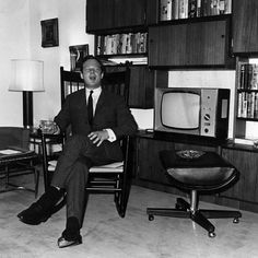 black and white photograph of man in suit sitting in chair with television on shelf behind him