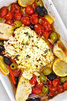 a white casserole dish filled with chicken, olives, tomatoes and bread