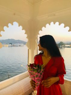 a woman in a red dress holding flowers looking out over the water from a gazebo