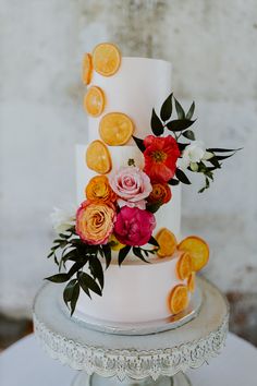 a three tiered white cake with orange slices and flowers on the top, sitting on a table