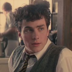 a young man with curly hair wearing a sweater vest and tie looking at the camera