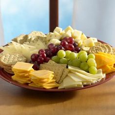 a plate full of cheese, crackers and grapes on a table next to a window