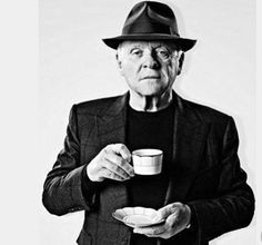 an old man holding a cup and saucer in his hands while wearing a hat