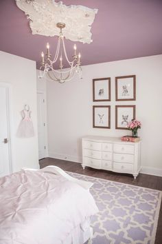 a white bed sitting under a chandelier next to a dresser in a bedroom