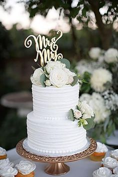 a wedding cake with white flowers and mr and mrs topper on the bottom tier