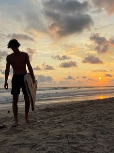 a man holding a surfboard on top of a beach next to the ocean at sunset