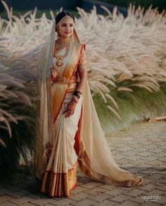 a woman in a white and orange sari standing next to some tall grass with her hands on her hips