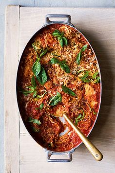 a pan filled with meat and sauce on top of a wooden table next to a spoon