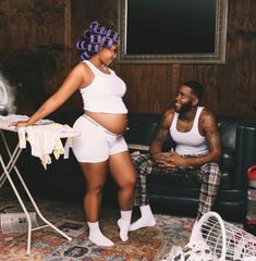 a pregnant woman standing next to a man sitting on a couch in a living room