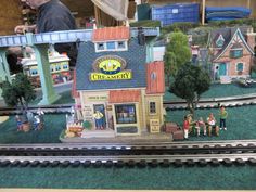 a toy train set with people on the tracks in front of a creamery shop