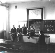 black and white photograph of men sitting at desks in front of chalkboard with writing on it