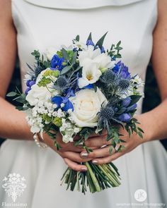 a bride holding a bouquet of white and blue flowers with greenery in her hands