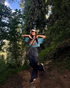 a woman standing on a dirt road in the woods with her arms behind her head