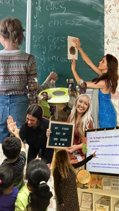 a group of children standing in front of a chalkboard with writing on it and two women holding up a sign