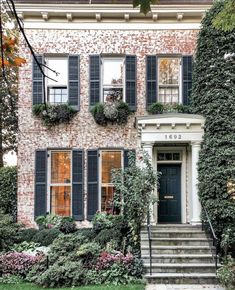 an old brick house with black shutters and green plants growing on the front door