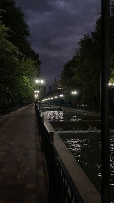 an empty walkway next to a body of water at night with street lights in the distance