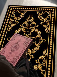 a person holding a pink book on top of a black and gold rug with an intricate design