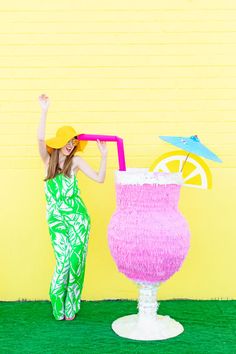 a woman standing next to a large pink drink in front of a yellow brick wall