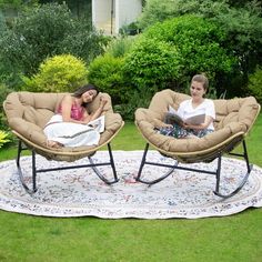 two people sitting in chairs reading books on a rug outside with trees and bushes behind them