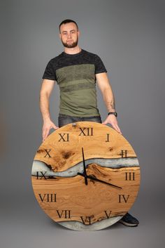 a man standing behind a large wooden clock with mountains and water on the face, in front of a gray background