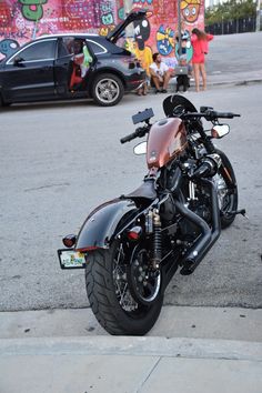 the motorcycle is parked on the side of the road with people standing around it and graffiti painted on the wall behind it